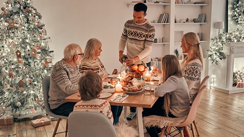 Imagem de uma família com seis pessoas, todas vestindo camisolas de lã bordadas, e todas à volta da mesa para uma refeição de Natal: um casal idoso, um rapaz e uma rapariga e um homem e uma mulher jovens. Estão todos sentados, com exceção do homem, que serve um copo com vinho. Ao seu redor, a decoração é composta por objetos e móveis brancos, prateados e em tons de madeira.