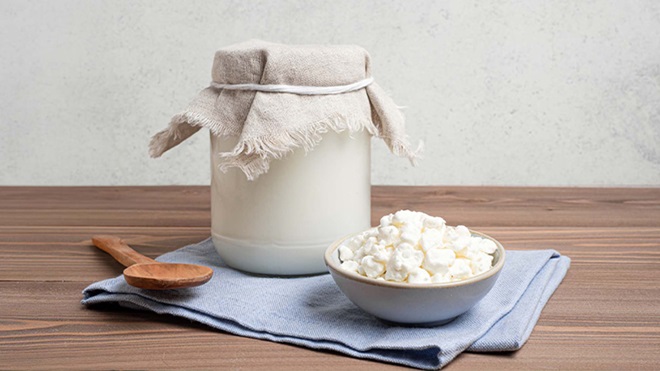 Frasco de iogurte coberto com pano sobre mesa de madeira, ao lado de tigela com kefir e colher de madeira, sobre guardanapo azul-claro.