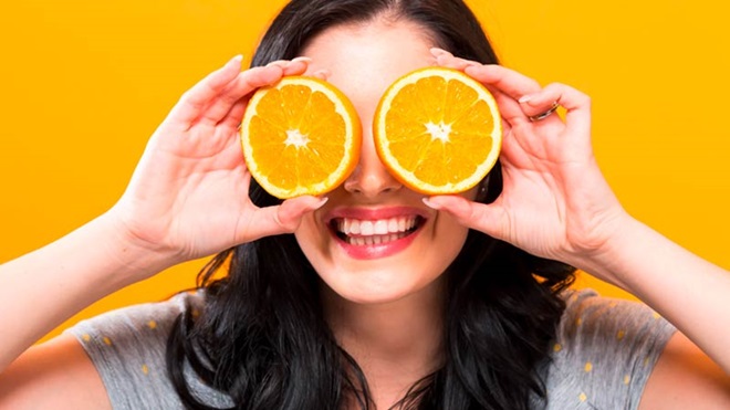 Mulher jovem segura duas metades de laranja à frente da cara, na zona dos olhos e sorri; fundo laranja - alusão a vitamina C