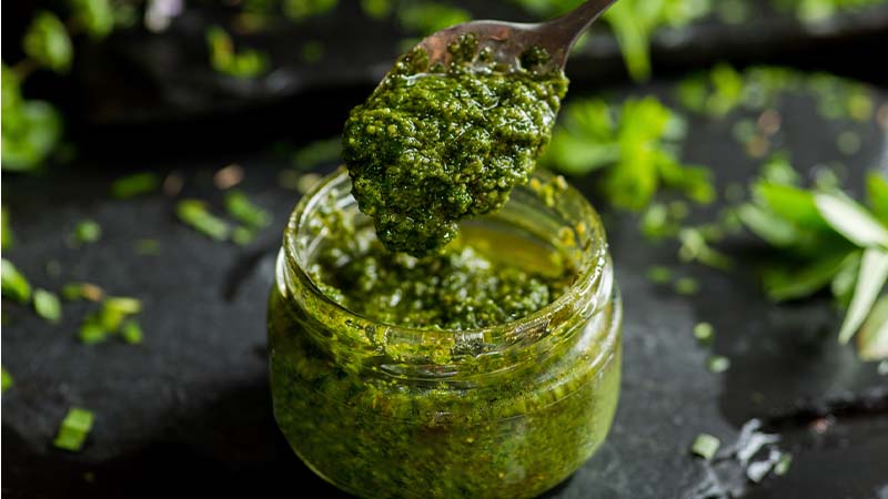 Frasco de vidro com molho verde espesso, tipo pesto, sendo servido com uma colher; ervas frescas e folhas verdes espalhadas sobre superfície escura.