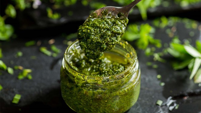 Frasco de vidro com molho verde espesso, tipo pesto, sendo servido com uma colher; ervas frescas e folhas verdes espalhadas sobre superfície escura.