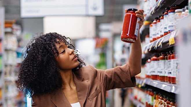 Pessoa num corredor de supermercado estende o braço para retirar um frasco de molho de tomate de uma prateleira, rodeada por estantes cheias de produtos.