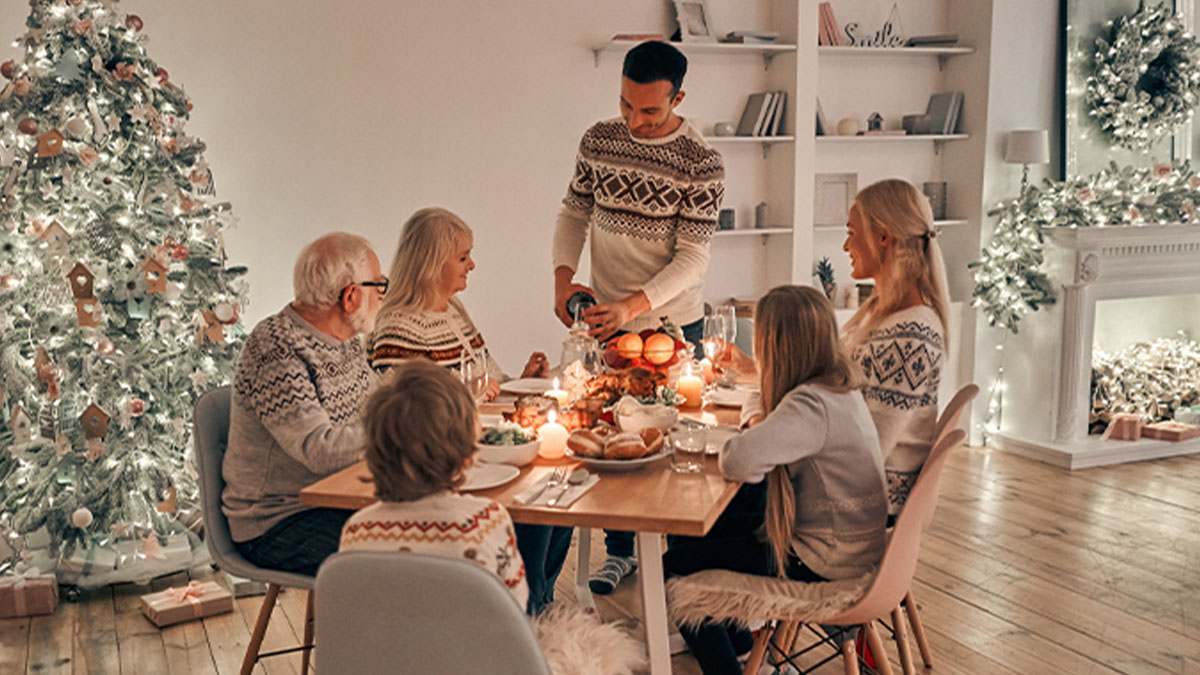 Família reunida à mesa de jantar decorada para o Natal, com árvore iluminada e lareira enfeitada ao fundo.