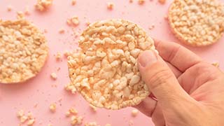 Mão a segurar bolacha de arroz ou milho tufado, fundo rosa com mais bolachas