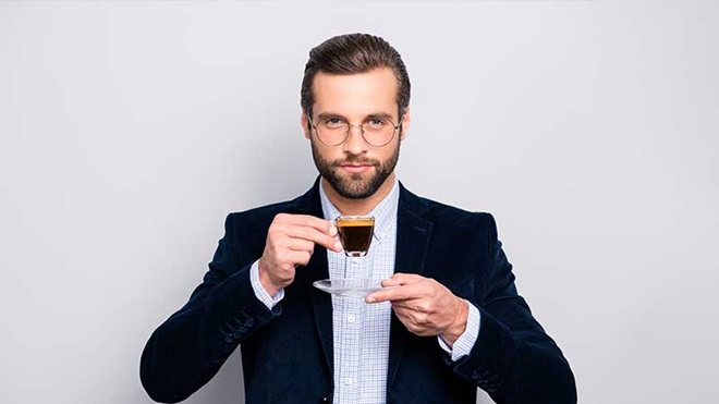 Homem de barba aparada, óculos, camisa e blazer a beber café expresso numa chávena transparente
