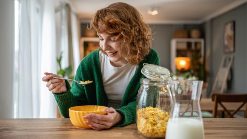 Mulher jovem come flocos de milho de uma taça, na sua cozinha