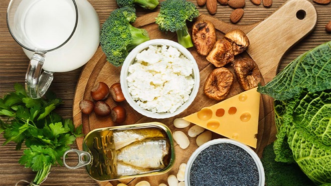 Mesa com alimentos ricos em cálcio: leite, queijo, iogurte, frutos secos, sementes, azeite, brócolos e folhas verdes, dispostos sobre madeira