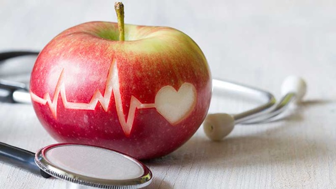 Maçã vermelha com gráfico de eletrocardiograma e coração por cima, envolta num estetoscópio, sobre uma superfície branca