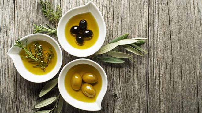 3 taças de louça branca em formato de lágrima sobre uma mesa de madeira: cada taça tem azeite, temperos e azeitonas - alusão ao azeite