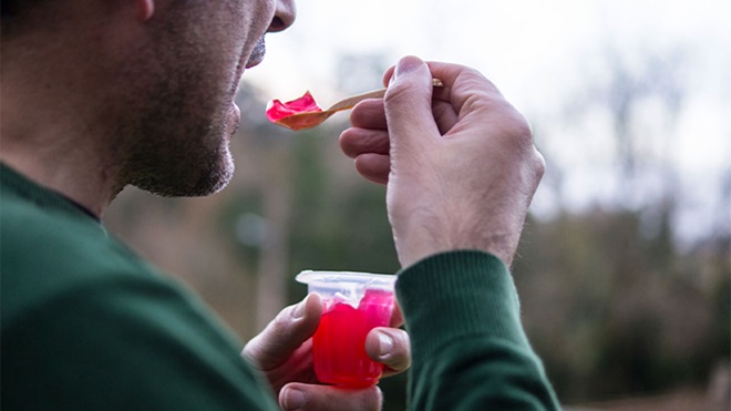 Homem com camisola verde escura a comer gelatina vermelha no exterior, árvores desfocadas ao fundo