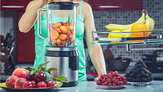 Pessoa a preparar fruta num liquidificador numa cozinha, com bananas, laranja, framboesas, amoras e maçãs sobre a bancada.