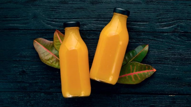 2 embalagens plásticas transparentes com sumo cor de laranja vivo e tampa preta, sobre uma mesa de madeira escura; folhas verde escuras por baixo das garrafas