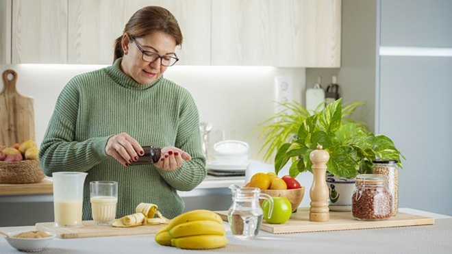 Mulher a tomar suplementos alimentares junto de uma bancada de cozinha com um copo de batido e fruta
