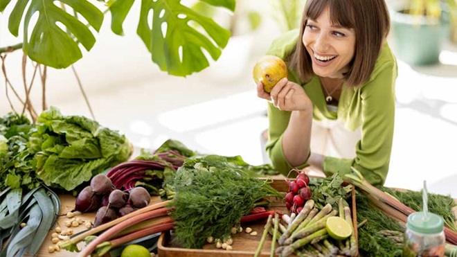 Mulher com ar sorridente pega num garfo à frente de uma mesa cheia de vegetais de várias cores porque pratica uma alimentação vegetariana