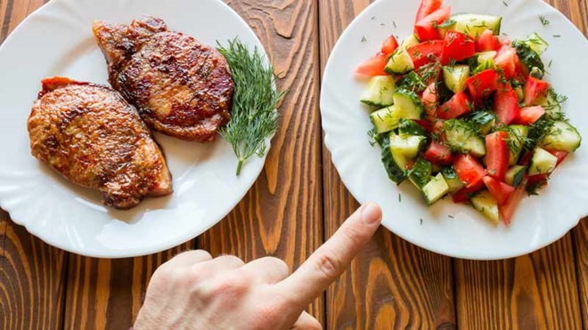 Dois pratos brancos em cima de uma mesa: um deles tem pedaços de carne grelhada e o outro uma salada. Uma mão aponta para o prato com a salada - alusão a Flexitarianismo
