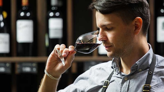 Sommelier cheira copo de vinho tinto frente a prateleiras com garrafas de vinho