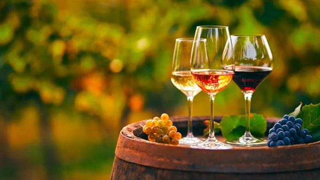 Três copos de vinho, tinto, rosé e branco, sobre uma pipa de vinho, com uvas ao lado e uma vinha ao fundo