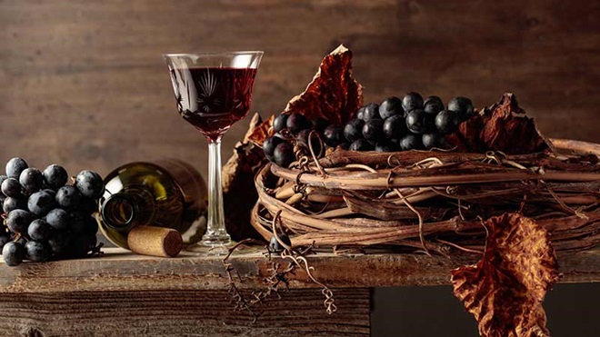 Cálice de vinho de Porto pousado sobre mesa de madeira ao lado de garrafa deitada, num cenário rústico com cacho de uvas e folha de videira seca