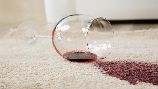 Copo de vidro com vinho tinto entornado no chão, numa carpete ceme; nódoa