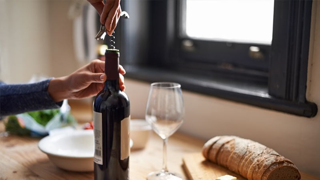 Mãos a abrir uma garrafa de vinho com saca-rolha, sobre uma mesa posta, com prato e copo de vinho