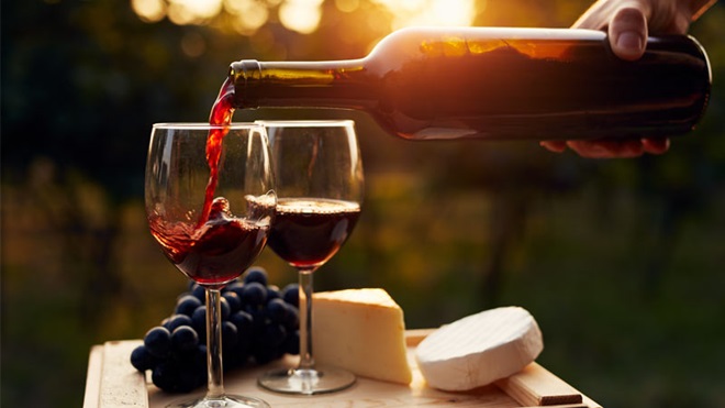 Pessoa a servir dois copos de vinho para acompanhar uma tábua de queijos decorada com um cacho de uvas