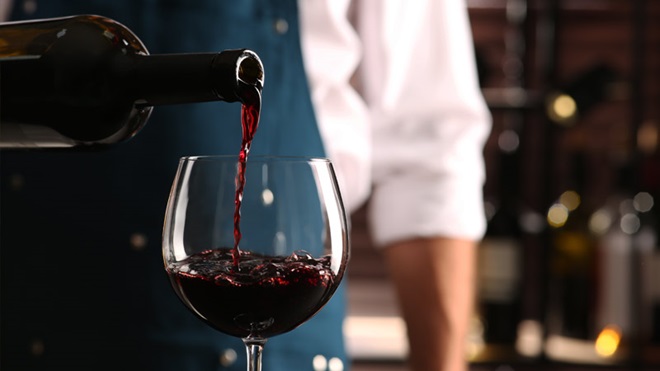 Grande plano de um homem com avental azul escuro a servir um vinho tinto num copo de vidro de balão