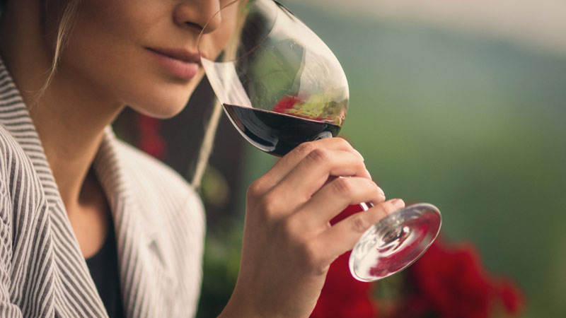 Grande plano de cara de mulher com casaco, a provar um vinho tinto num copo de vidro alto