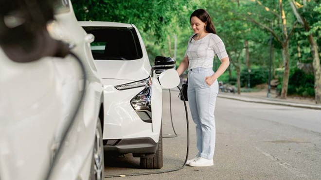 Mulher jovem carrega um carro elétrico branco no exterior, com árvores ao fundo