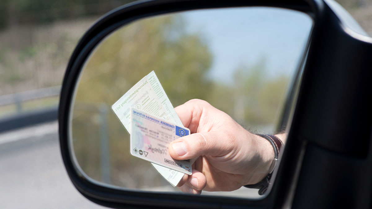 Grande plano de um espelho onde se vê um condutor a mão de um condutor a segurar os documentos do carro e carta