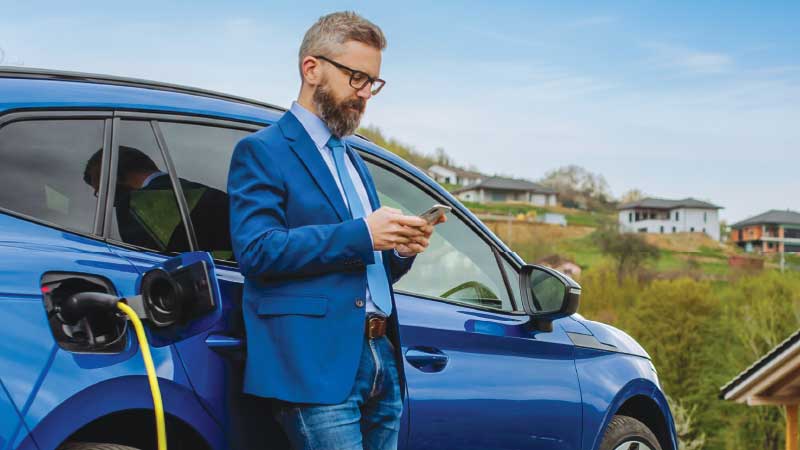Homem ao telemóvel encostado ao carro elétrico azul enquanto carrega na rede pública