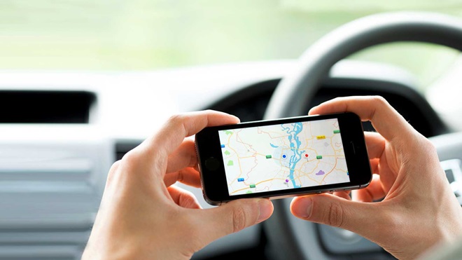 Imagem no interior de um carro, com o tablier e volante como fundo, e em primeiro plano as mãos de um condutor segurando um telemóvel com uma app de GPS visível no ecrã.
