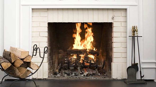 Lareira a lenha acesa, ferramentas de mexer o lume do lado direito; pequena pilha de lenha arrumada num suporte do lado esquerdo. Parede branca à volta.