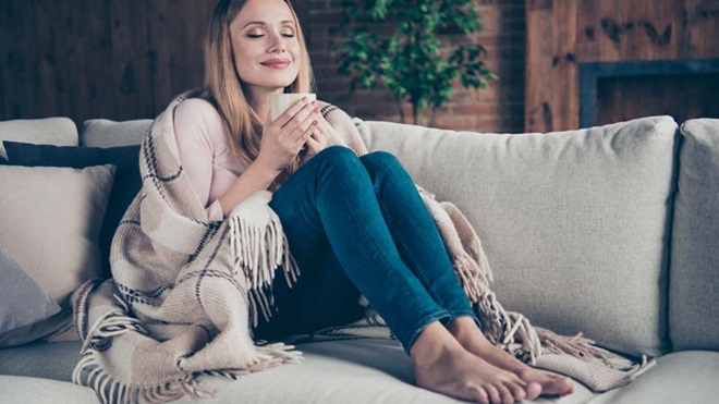 Mulher confortável em casa, sentada no sofá com uma manta a beber um chá