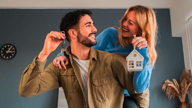 Casal jovem dentro de uma casa, segurando uma casinha de bricar e sorri