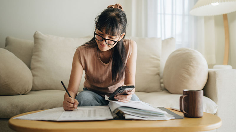Mulher sentada no sofá a trabalhar numa mesa baixa, escrevendo em papéis com uma caneta enquanto segura um telemóvel; ao lado, uma caneca e vários documentos espalhados.