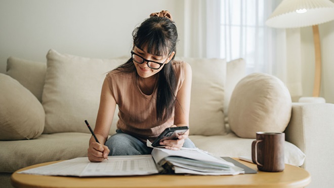 Mulher sentada no sofá a trabalhar numa mesa baixa, escrevendo em papéis com uma caneta enquanto segura um telemóvel; ao lado, uma caneca e vários documentos espalhados.