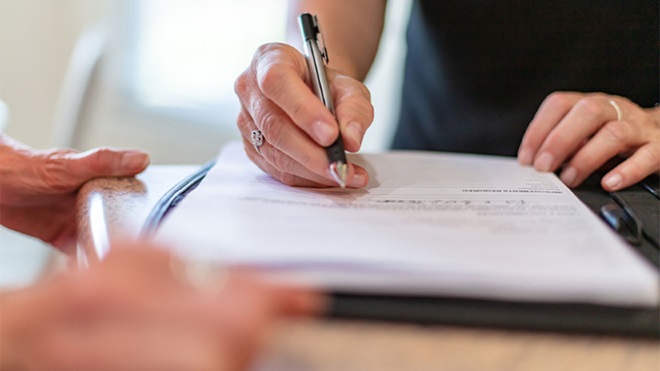 Pessoa a assinar um documento sobre uma mesa, segurando uma caneta enquanto outra pessoa observa do lado.