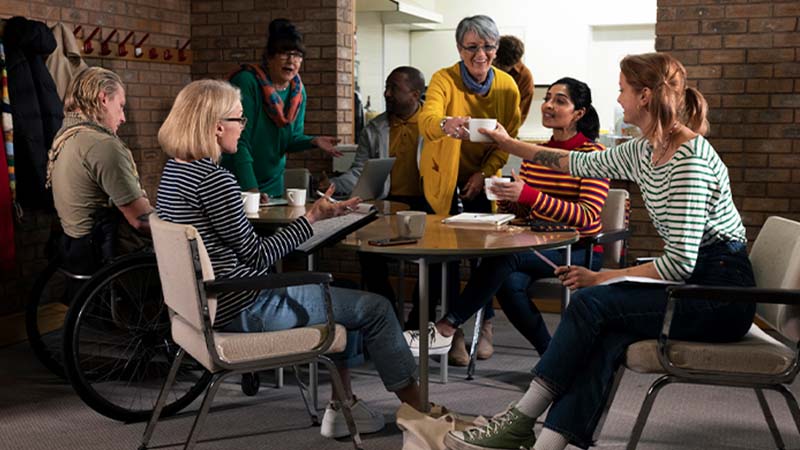 Grupo de pessoas a conversar e partilhar bebidas numa sala comunitária, sentadas e de pé em torno de uma mesa, num ambiente acolhedor e descontraído.