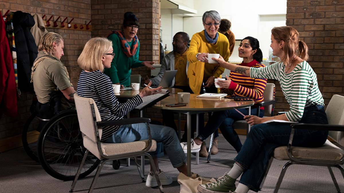 Grupo de pessoas a conversar e partilhar bebidas numa sala comunitária, sentadas e de pé em torno de uma mesa, num ambiente acolhedor e descontraído.