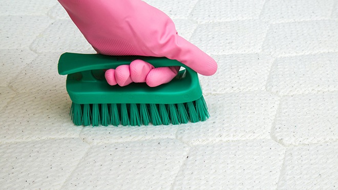 mão com luva cor de rosa a limpar colchão com escova verde
