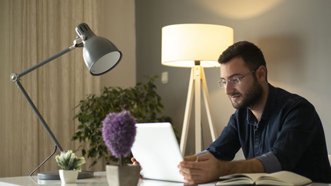 Consumo de eletricidade em casa - homem sentado no escritório em casa, ao computador, com dois candeeiros próximos, um ligado e outro desligado 