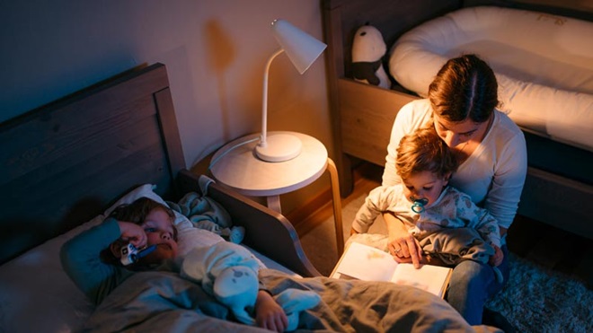 Mãe com dois filhos pequenos lê uma história no quarto à hora de deitar; candeeiro ligado