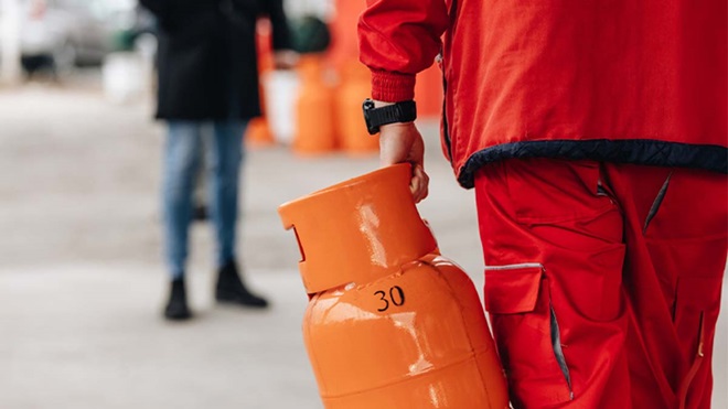 Pessoa com uniforme vermelho transporta uma botija de gás laranja marcada “30” numa área exterior, com outra pessoa desfocada e elementos industriais no fundo.