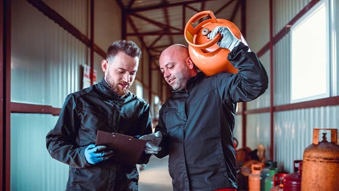 Dois técnicos de gás analisam um relatório dentro de um armazém; um deles carrega uma botija de gás laranja ao ombro