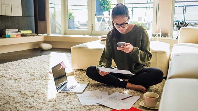 mulher sentada no chão de uma sala, com o computador ao lado e papéis, a resolver problemas de faturação no telemóvel.