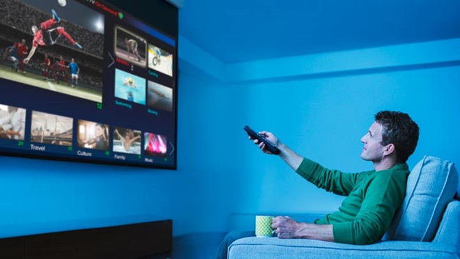 Homem adulto sentado no sofá a ver televisão com o braço esticado a segurar e a apontar o comando