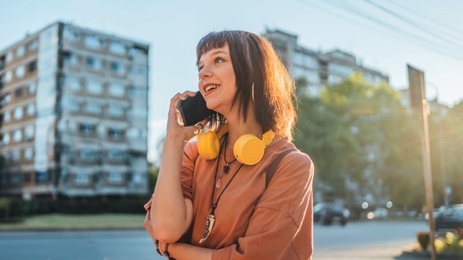 Jovem numa rua da cidade, com headphones no pescoço, fala ao telemóvel