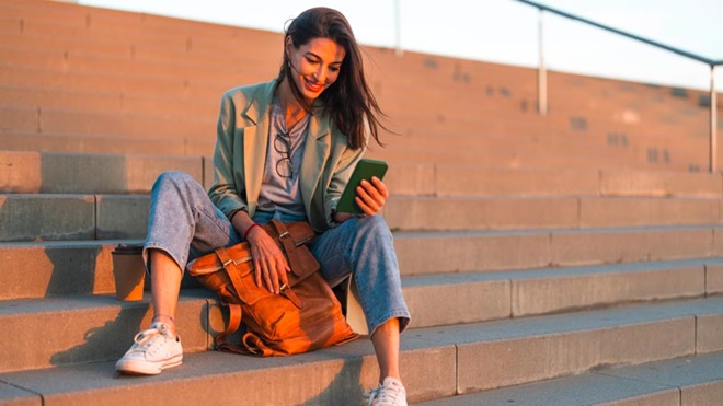 Mulher jovem sentada numas escadas no exterior, olha para o ecrã de um telemóvel