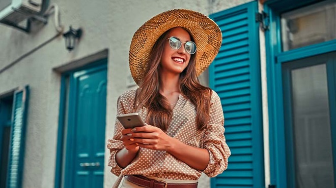 Mulher sorridente com chapéu e telemóvel na mão, de férias no exterior de uma rua histórica, numa cidade europeia