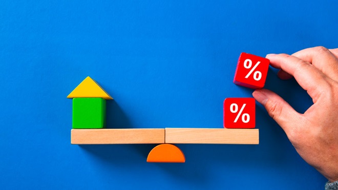 Peças de madeira sobre uma mesa formando uma balança com uma casinha de um lado e cubos com percentagens do outro